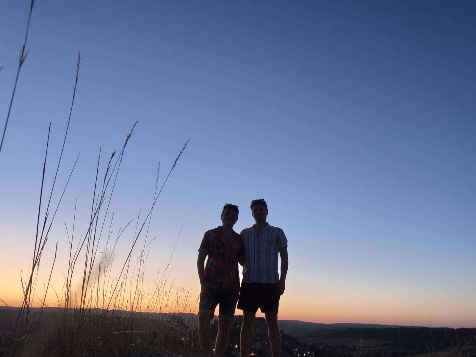 Ale and Harry on a grassy hilltop at dusk, the sky orange along the horizon and the valley below them.